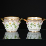 <b>Par de "cachepots" com alças</b>, em porcelana dinamarquesa "Royal Copenhagen", modelo <b>"Flora Danica"</b>, profusamente decorados em policromia com ramagens e botões de flores em relevo e bordas douradas. Cada qual com sua identificação botânica no verso. Alt. 12 cm; Diâm. 21 cm
