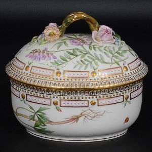 <b>"Bombonnière" com tampa</b> em porcelana dinamarquesa da importante manufatura Royal Copenhagen, modelo <b>"Flora Danica"</b>. Peça inteiramente pintada à mão em belíssima policromia com suas designações botânicas e numeração no verso. Apresenta borda em mínimos picotados cobertos a folha de ouro. Compr. 15,5 cm