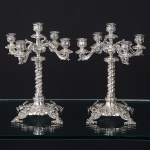 <b>Par de candelabros para 5 luzes</b> em prata portuguesa. Base quadrada profusamente repuxada e cinzelada com flores e "coquilles". Pés projetados em elaboradas composições vegetais. Haste torcida apoiando 4 braços movimentados recamados por volutas. Contrastes portugueses. Alt. 32,5 cm. Peso 4600 gramas.