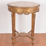 <b>Mesa </b>circular no estilo Louis XVI em madeira patinada a ouro e entalhada com goivos e guirlandas. Pernas retas decoradas com frisos escavados terminados em ponteiras. Tampo em alabastro. Mínimos descascados no ouro. Alt. 78 cm; Diâm. 64 cm