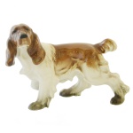 Estatueta em porcelana alemã, policromada representando cachorro hound. No fundo marca da manufatura. 10 x 14 cm.