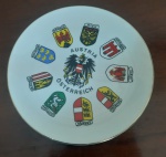 Prato decorativo em porcelana KM Áustria (Österreich) com brasão ao centro e bandeiras estatais. Esta peça de souvenir é ideal para colecionadores, destacando-se por seu design elegante - Diâmetro: 19 cm