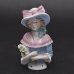 Coleção Half Doll - dama "Florista", com sua elegante roupa de campo, chapeu de largas abas com grande laço cor de rosa, corpete azul de gola rosa com binando com laço, carrega em uma das mãos buque de flores do campo. Base numerada.  Mede 11x6 cm