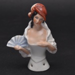 Coleção Half Doll - Jovem dama segura leque em uma das mãos e apoia as outras no seio. Vestimenta toda branca com grande decote. Chapéu na cor vermelho com plumas. Base numerada e marcada Germany. Med 9x7 cm.