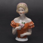 Coleção Half Doll - Jovem dama com vestido de babados laranja que deixa o seu colo aparente. Segura uma rosa em uma das mãos e tem cabelos chnel graciosos e curtos. Base numerada. Mede 10x7 cm.