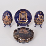 Blue & Gold - Lote composto por conjuto de peças em porcelana em tons de azul cobalto e detalhes em ouro, sendo: 1 Medalhão miniatura "Miranda de Ebro" , 1 porta jóias Limoges, tampa com cena galante e, 2 ovos estilo Fabergè com decoração floral - Total 4 peças. Obs.: Porta joias com parte colada no fundo e 1 ovo sem a presilha, no estado. (Diam 10cm / 4x10 / 10x6)