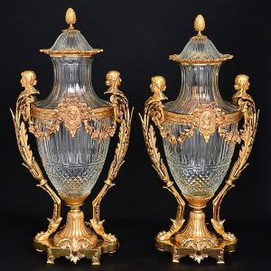 <b>MAISON BACCARAT - Magnífico par de ânforas </b>em cristal francês BACCARAT translucido e multi lapidado, com montagem em bronze ormolu. Duplas alças em cariátides implantadas em folhas e unidas em guirlandas centralizando camafeus perolados, encimados por laços. Pés em garras. Base em pedestal recortado decorado com volutas. Pegas das tampas modeladas em pinhas. Séc. XIX. Marca da "cristallerie" no bronze e no cristal. Alt. 73 cm