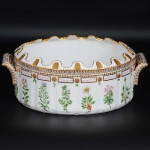 Excepcional <b>"rafraîchissoir" </b>oval de borda ondulada em porcelana dinamarquesa da importante manufatura Royal Copenhagen modelo "Flora Danica". Inteiramente pintada à mão em belíssima policromia com suas designações botânicas e numeração no verso. Apresenta borda em mínimos picotados cobertos a folha de ouro. Med. 33 x 23 cm