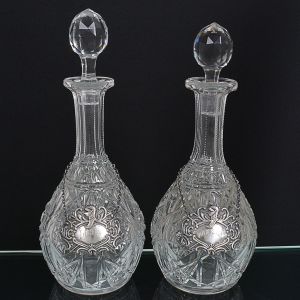 <b>Par de garrafas </b>piriformes em cristal francês BACCARAT profusamente lapidado em estilizações geometrizadas. Hastes serrilhadas e tampas multifacetadas. Marca da "cristallerie" na base. Acompanha colares em prata brasileira 833 ml. Interior das tampas com bicado na parte interna. Alt. 32 cm