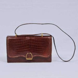 <b>HERMÈS</b> - Paris - Antiga <b>bolsa</b> em couro de crocodilo na cor marrom com ferragens douradas, tendo o fecho forma de estribo. Med. 26 x 17 x 6,5