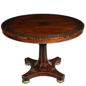 Bela <b>mesa de apresentação</b> inglesa, "Regency", de forma circular, construída em pluma de mogno. Decoração da borda com embutidos metálicos formando arabescos vegetais. Montante central facetado sobre base em arcada. Pés metálicos em bolas chapadas. Séc. XIX. Alt. 78 cm; Diâm. 122 cm