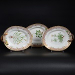 <b>Conjunto de 3 travessas ovais com alças</b>, em porcelana dinamarquesa da importante manufatura Royal Copenhagen modelo "Flora Danica". Cada peça, inteiramente pintada à mão em belíssima policromia com suas designações botânicas e numeração no verso. Apresenta borda em mínimos picotados cobertos a folha de ouro. Med. 25 x 20 cm