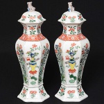 <b>Par de potiches </b>em porcelana Companhia das Índias, apresentando forma alongada com bojo em facetados decorados com vasos e composições floridas predominando as tonalidades de verde, azul e "rouge de fer". Tampas encimadas por cães de fó. Período Kangxi (1662-1722). Alt. 33 cm