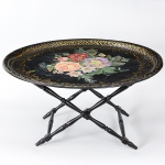 Bela <b>bandeja inglesa</b>, vitoriana, de forma oval, em latão negro, pintado com arranjo de flores em policromia e arabescos a ouro. Possui assinatura. Acompanha tripé em madeira ebanizada. Med. 76 x 61 cm