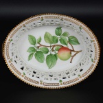 <b>Fruteira </b>oval em porcelana dinamarquesa da importante manufatura Royal Copenhagen modelo "Flora Danica". Peça inteiramente pintada à mão em belíssima policromia com suas designações botânicas e numeração no verso. Apresenta borda em mínimos picotados cobertos a folha de ouro. Med. 23 x 18,5 cm