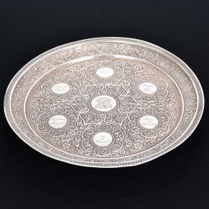 <b>Bandeja </b>circular em prata, europeia, profusamente decorada, com centro lavrado com o brasão do estado da Guanabara cercado por medalhões com inscrições datadas de 1904. Restante da peça com profusa decoração de flores. Borda perolada. Pés esféricos (faltando um). Peso 600 g. Diâm. 30,5 cm