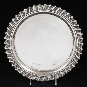 <b>Bandeja</b> circular em prata portuguesa. Apresenta centro liso e borda em gomos levemente oblíquos. Contraste da cidade do Porto usado a partir de 1938. Teor 833 ml. Peso 485 g. Diâm. 29 cm