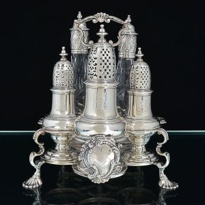 Raro <b>galheteiro "cruet stand"</b> em prata inglesa do séc. XVIII apresentando suporte com alça central decorada com volutas e "crest".  Em uma das faces belo escudo armorial cercado por volutas e pequenos frutos. Pés concheados. Apresenta três polvilhadores "casters" e dois recipientes em cristal lapidado. Contraste da cidade de Londres de 1743. Período George II. Alt. 24,5 cm