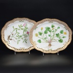 <b>Par de travessas</b> ovais com bordas reticuladas em porcelana dinamarquesa da importante manufatura Royal Copenhagen modelo "Flora Danica". Cada peça, inteiramente pintada à mão em belíssima policromia com suas designações botânicas e numeração no verso. Apresenta borda em mínimos picotados cobertos a folha de ouro. Med. 27 x 22,5 cm