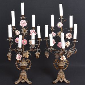 <b>Par de candelabros</b> europeus com estrutura em metal com vestígios de dourado. Decoração com cachos de uvas, folhas de parreira e variadas flores e velas em porcelana. Base trípode. Uma vela colada. Adaptados para luz elétrica. Séc. XIX. Alt. 85 cm