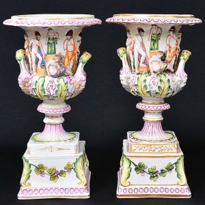 <b>Par de ânforas </b>em porcelana italiana Capo di Monti, decoradas em relevo e policromia com figuras da antiguidade romana. Alças implantadas em cabeças antropomorfas. Base quadrangular decorada com guirlandas. Marca da manufatura no verso. Séc. XIX. Alt. 34,5 cm