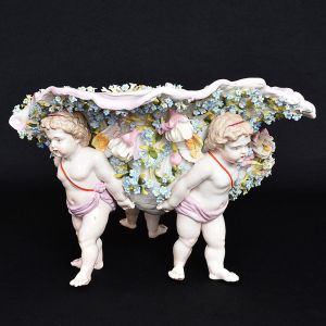 <b>Centro de mesa </b>em porcelana Meissen em forma de "coquille", decorado com profusão de flores em policromia, sustentado por três figuras angelicais. Marca da manufatura e numeração no verso. Compr. 36 cm; Alt. 23 cm