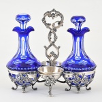 <b>Galheteiro</b> com montagem em prata francesa, apresentando alça central e suportes decorados em vazados com volutas, flores e pequenas uvinhas. Pés revirados. As galhetas em cristal BACCARAT translúcido com "overlay" em azul cobalto e lapidação com trevos e geometrizados. A prata com contraste do séc. XIX. Falta um pezinho do centro. Peso 480 g. Alt. 21 cm