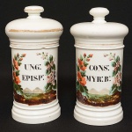 <b>Par de potes de farmácia </b>em porcelana francesa branca, apresentando a designação dos produtos (CONS: MYR: B: e UNG: EPISP) cercada por policromia de flores, folhas e montanhas. Filetes a ouro com desgastes. Um deles com pequeno bicado e o outro com tampa defeituosa e pequeno bicado na parte interna. Alt. 28 cm