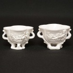 <b>Par de taças de libação </b>em porcelana chinesa, "blanc de Chine", oblongas e quadrilobadas. Bojos ornamentados por ramos de cerejeiras em flor, aplicados em alto relevo até as bases. Alças laterais em argolas. Séc. XVIII. Alt. 6,5 cm