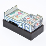 Raro e delicado <b>descanso de pinceis</b>, em porcelana chinesa policromada com realces a ouro. Apresenta formato retangular com extremidades recortadas em arcos e frente com degraus, ornamentada por cena com dama e atendente em jardim, ladeada por composições florais. Laterais vazadas. Sob a base, marca do Reinado Daoguang (1821-1850). Acompanha peanha em madeira laqueada. Med. 8 x 4,5 cm