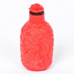 "<b>Snuff bottle</b>" retangular, chinesa, em pasta de vidro de cor coral, profusamente esculpida com dragões perseguindo pérola no meio de nuvens. Tampa no formato de flor de mesma cor, com colar preto e colher. Séc. XIX. Alt. 8 cm