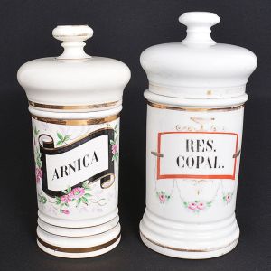 <b>Conjunto de 2 potes de farmácia</b>. Um em cerâmica creme com rótulo e policromia florida e frisos a ouro e craquelado e com discretos bicados na borda do pote. Outro em porcelana branca com rótulo em policromia de guirlandas e imperceptível bicado na borda da tampa. Alt. 26 cm e 28 cm