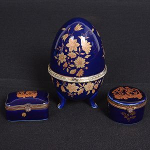 <b>Conjunto de três caixas </b>em porcelana azul noite, as duas menores decoradas com cenas galantes e marca da manufatura de Limoges. Outra maior em forma de ovo, com montagem metálica (montagem necessitando colar). Alt. da maior 13 cm