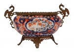 Arte Oriental - Belíssima e rara floreira no formato oblongo em gomos de porcelana japonesa dita "Imari", profusamente decorada, em sua policromia característica, pelos esmaltes nas cores azul índigo, verde, vermelho e "rouge de fer" sobre fundo branco. Em reserva paisagem de jardim com ponte.  Japão -Séc. XIX/ XX. Med. 22 x 37 x 18 cm (ME). Marcas do tempo. Presença de defeito de forno e fio de cabelo no fundo.  Apresenta guarnição de metal ao gosto do estilo império, com pegas na forma de grifos que fora adaptada posteriormente. Esta guarnição está em mau estado de conservação.  Coleção Particular Rio de Janeiro/RJ. Nota: PORCELANA JAPONESA - IMARI. Das muitas escolas consagradas de cerâmicas e porcelanas japonesas, possivelmente a Imari é das mais conhecidas no exterior por estar sendo exportada desde o final do século XVII, ainda quando o relacionamento internacional do Japão era efetuado exclusivamente pelos holandeses, que passou por uma abertura na Era Meiji. O acervo mais importante de peças Imari encontra-se atualmente no Museu de Cerâmica Oriental de Osaka. Como é sabido, os japoneses aperfeiçoaram as técnicas aprendidas dos chineses, por intermédio dos artistas coreanos, tendo se concentrado inicialmente na região sul do Japão, mais próxima da península coreana. Sua exportação substituiu a chinesa, sendo efetuada pelo porto Imari, ficando consagrada por este nome. Os chineses já produziam porcelanas quando a Europa ainda contava somente com cerâmicas de baixa temperatura, como estão apresentadas no Museu de Servrés, na França. As peças chinesas ficaram difíceis de serem exportadas para a Europa diante dos distúrbios políticos na passagem da dinastia Ming para a Qing, na década de 1640. A Companhia das Índias Orientais holandesa tinham um entreposto em Nagasaki e a exportação do Japão começou em 1659. Estas peças de porcelana eram adequadas para os palácios e residências da aristocracia europeia. As grandes peças Imari eram utilizadas como ornamentos e aparentam pouco uso. Mas existem variadas peças, como pratos e outros utensílios, todos com desenhos característicos, sendo identificados também pelos esmaltes utilizados. É comum encontrar ainda hoje peças de séculos nos antiquários de todo o mundo, que atualmente alcanças preços elevados. A utilização do ouro, que já atraía os europeus desde a época de Marco Polo, mas que surpreendem os bons apreciadores de cerâmicas e porcelanas, sendo que existem escolas que mantiveram seus estilos, principalmente na região de Arita, entre os quais um bastante conhecido é o chamado Kakiemon. Tudo indica que muitas destas peças eram encomendadas pelos europeus, onde algumas cores que eram obtidas pelos produtores japoneses também se destacavam, permitindo o desenvolvimento do chamado Japonismo no século XIX. Nesta época, a admiração era recíproca e também os japoneses se interessavam pelas obras de arte, notadamente dos artistas europeus, com destaque dos franceses. Fonte: https://criart.com.br/wp-content/uploads/2021/03/Material-Extra-Porcelana-Japonesa.pdf