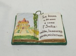 Placa decorativa na forma de livro, descrevendo idades da mulher em italiano, em cerâmica. Medindo 14cm x 10cm. Leve bicado.