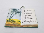 Placa decorativa na forma de livro, descrevendo idades da mulher em italiano, em cerâmica. Medindo 14cm x 10cm. Leve bicado.