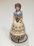 Delicado sino em porcelana no formato de camponesa, ricamente decorado com pintura floral policromada e acabamento vitrificado. A figura apresenta lenço vinho com pintas brancas, echarpe azul xadrez e vestido em tom creme com belos detalhes campestres, segurando um pequeno buquê junto ao corpo.Peça encantadora, de inspiração europeia, ideal para colecionadores e decoração temática. Mede aproximadamente 17 cm de altura e acompanha suporte metálico para exposição.