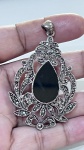 PINGENTE EM METAL PRATEADO COM PEDRAS E PEDRA CENTRAL EM CABOCHÃOPingente em metal de tonalidade prateada, com estrutura rendilhada de inspiração clássica, ricamente trabalhada com aplicações de pequenas pedras facetadas, formando composição ornamental detalhada em padrão floral e arabescos.Elemento central composto por pedra em formato de gota (cabochão), em tonalidade escura, com superfície lisa e polida, proporcionando contraste elegante com as demais aplicações.Argola superior integrada à peça, permitindo uso em correntes diversas.PERÍODO: contemporâneo com inspiração vintageMATERIAL: metal prateado e pedras (não identificadas)TÉCNICA: fundição, cravação e polimentoESTADO DE CONSERVAÇÃO: bom estado geral, com leves sinais de uso compatíveis, sem perdas aparentes nas aplicações.OBSERVAÇÕES: peça de estética sofisticada, com forte influência vintage, destacando-se pela riqueza de detalhes e pelo contraste entre a pedra central e as demais aplicações brilhantes. Ideal para uso ou composição de conjunto com acessórios de estilo clássico