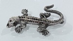 BROCHE EM PRATA DE LEI 925 COM MARCASSITAS EM FORMATO DE LAGARTOBroche confeccionado em prata de lei 925, representando figura de lagarto com rica composição de detalhes em relevo.A peça é inteiramente adornada com marcassitas aplicadas, distribuídas ao longo do corpo, cauda, cabeça e membros, criando efeito de brilho discreto e sofisticado. Algumas pedras apresentam tonalidade mais escura, contribuindo para contraste e profundidade visual.Modelagem tridimensional bem definida, com acabamento minucioso que valoriza as escamas, patas e expressão da peça. Cauda sinuosa e composição dinâmica reforçam o caráter decorativo e expressivo do broche.Trata-se de peça antiga, de caráter colecionável, com forte apelo ornamental e inspiração naturalista. Fecho posterior funcional.