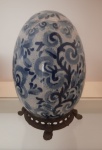 Elegante ovo em porcelana craquelê oriental é um exemplar distinto de arte e sofisticação. Ornamentado com delicadas flores em tom de azul, ele exibe um acabamento sofisticado que realça sua beleza. A peça é acompanhada por uma base de madeira refinada, que complementa o design e oferece um suporte ideal para exibição. Altura: 18 cm