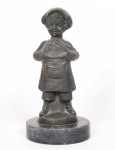 ESCULTURA em bronze patinado representando um poulbot, moleque pobre de Montmartre em Paris, que ganha a vida prestando pequenos serviços, de pé sobre base circular. Exibe o tradicional avental de peito de mesmo nome, o poulbot. Peanha em mármore preto. Med. Alt. 20,0 cm. Diâm. 10,0 cm. (base).