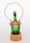 GALHETEIRO para azeite em forma de lampião com caixa de música com chave sob a base para  dar corda com estrutura em cobre vermelho. Bojo circular em vidro verde, no formato de garrafa, apoiado em base lisa. Hastes laterais em torsades, sustentando cúpula abobadada com alça superior arqueada. Parte central encimada por gargalo longo e adorno cônico. Med. Alt. 22,0 cm  Diâm. 10,0 cm.