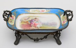 SÉVRES - Antiga fruteira em porcelana francesa de Sévres, com aplicações em bronze com guirlandas de flores, alças em forma de folhagens revirada. No interior cena romântica com jovem ensinando a sua amada tocar flauta. Marca da manufatura no verso. Séc. XIX.  Pequeno restauro antigo.  Med. Alt. 15,0 cm  Larg. 18,0 cm   Compr. 32,0 cm
