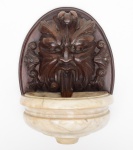 FONTE DE PAREDE - Pequena e antiga fonte de parede construída na parte superior com rosto masculino em madeira  finamente entalhada com orifício de saída da água e parte inferior com  bacia em mármore de carrara. Perdas de entalhes. Med. Alt. 33,0 cm  Larg. 25,0 cm.
