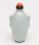 SNUFF BOTTLE de formato ovalado, em porcelanachinesa revestida por coberta monocromática celadon do tipo Ge com finarede de craquelê, esta mais escura de um lado da peça. Bojo abaulado,ladeado por pegas estilizadas. Tampa com colar em bronze, cabochão de corcoral e colher. Séc. XIX.