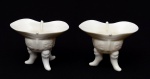 COLECIONISMO - Raro par de taças de libação em porcelana chinesa Blanc de Chine. Bojos globulares, moldados em alto relevo com reservas e ladeados por alças no formato de dragão. Bordas oblongas e reviradas, realçadas por frisos em gregas e encimadas ao centro por pequenas ponteiras. Pernas recurvas apoiadas em máscaras. Séc. XVIII. Peças de coleção. Med. Alt. 8,0 cm Larg. 11,0 cm.