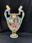 Faenza Italy.Grande vaso em fina maiólica de faiança FAENZA ITALY do início do seculo XX, com uma cena pintada no centro. A cena representa o mito de Perseu e Andrômeda. Perseu é visto libertando Andrômeda, que está acorrentada a uma fonte, enquanto uma figura masculina mais velha observa. O vaso possui duas alças laterais com figuras esculpidas e uma borda superior decorada com um rosto. As cores predominantes são azúis, amarelo, verde e tons de rosa e bege para as figuras. A base do vaso também é decorada com padrões geométricos e florais. A maiólica é um tipo de cerâmica italiana, conhecida por seus esmaltes brilhantes e cores vibrantes.Revisar as fotos atentamente, contem dois pequenos picados na base. Como se encontra nas fotos, medindo 55 cm de altura. 