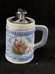 Vintage isqueiro de mesa antigo que se assemelha a uma caneca de cerveja alemã tradicional, também conhecida como stein. O corpo do isqueiro é feito de cerâmica branca com detalhes em azul e dourado, e apresenta uma pintura de um casal em cena galante com trajes de época. A parte superior do isqueiro, onde se encontra o mecanismo, em metal cromado. Como se encontra nas fotos medindo 13 cm de altura.