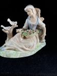Tengra.Bela escultura de fina porcelana espanhola, Tengra, que é conhecida pela sua alta qualidade e detalhes delicados. A peça retrata uma jovem sentada, segurando uma cesta de flores, com dois pássaros pousados em um tronco ao seu lado. As cores são suaves, predominando tons de branco, rosa e azul claro, o que confere à peça um ar delicado e romântico. A base da estatueta simula um terreno gramado, com nuances de verde e branco.Em ótimo estado de conservação, como se pode visualizar nas fotos, medindo 24 cm de comprimento por 21 cm de altura.   