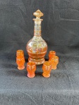conjunto de uma garrafa e seis copos de vidro "Carnival Glass  Jacobean  Josef Inwald". Este tipo de vidro é conhecido por seu brilho iridescente, que se assemelha ao efeito de óleo na água. O conjunto é composto por uma garrafa com tampa e seis copos pequenos. O vidro tem uma cor âmbar com reflexos iridescentes, e o design apresenta um padrão geométrico que lembra o estilo Jacobean. Em bom estado de conservação como se pode observar nas fotos, a garrafa mede 26 cm de altura.  